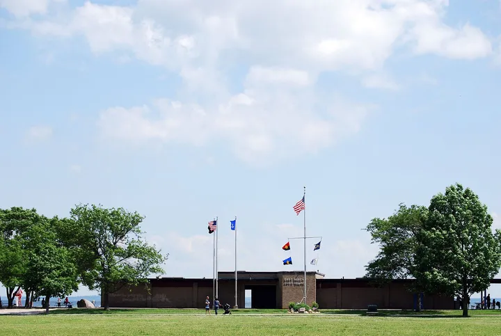 The East Beach building at Cove Island Park, a long, low brick pavilion with flagpoles, behind a wide lawn and shade trees, with Long Island Sound visible at the edges.