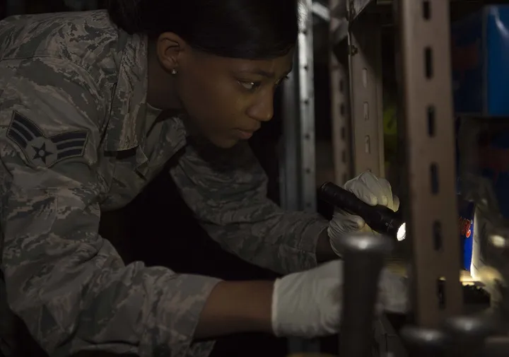 A pest management technician in gloves shines a flashlight into the back of a storage shelf.