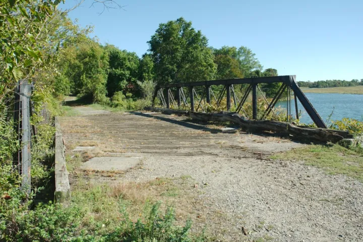 An old steel truss bridge with a worn plank deck on a gravel path in Pine Creek Park, with trees on the left and the open water and marsh of Pine Creek on the right.